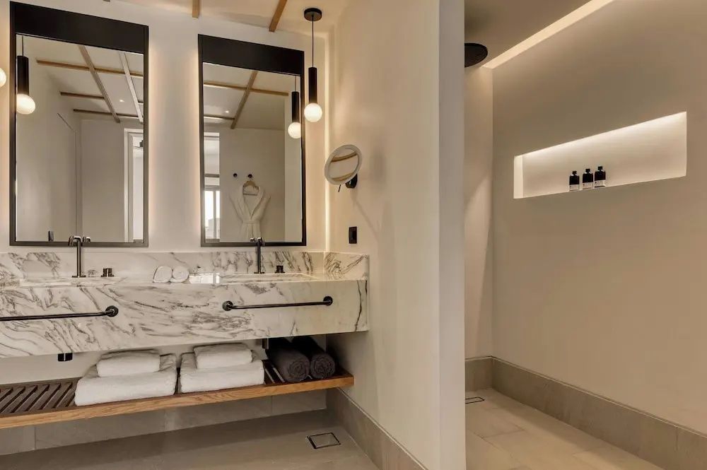 Badezimmer mit hellem Marmor-Waschtisch, Spiegeln, Handtüchern und abgetrenntem Duschbereich.