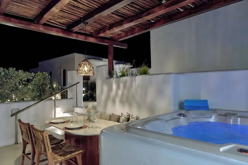 Beleuchtete Terrasse am Abend mit Whirlpool, Esstisch und Sicht auf weiße Gebäude.