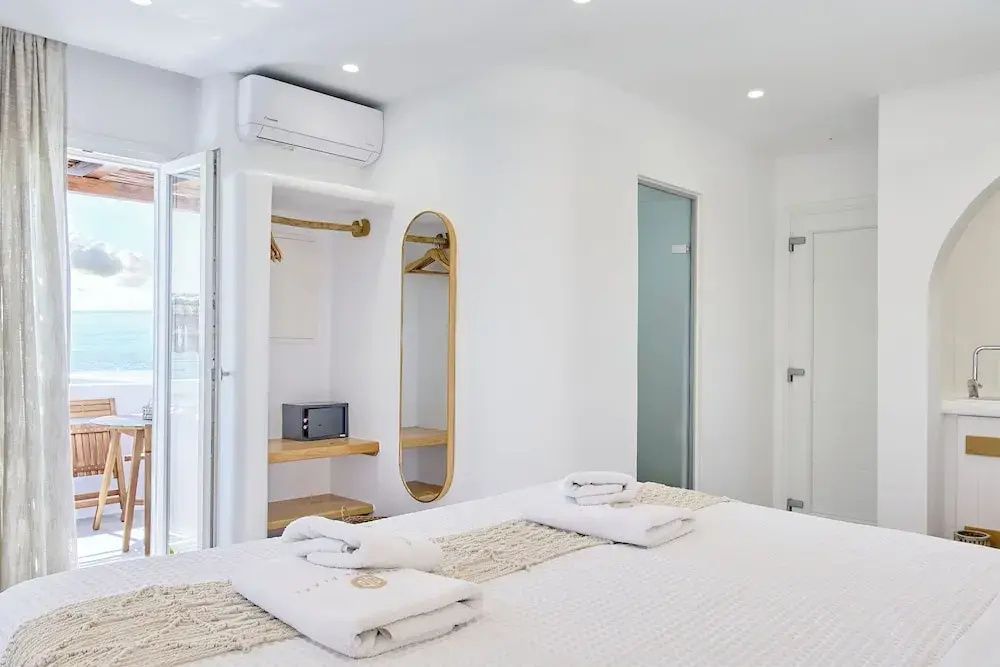 Weiß eingerichtetes Hotelzimmer mit offenem Kleiderschrank, Wandspiegel und Safe.