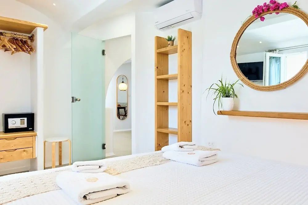 Helles Hotelzimmer mit Holzregal, rundem Wandspiegel und Glastür zum Bad.