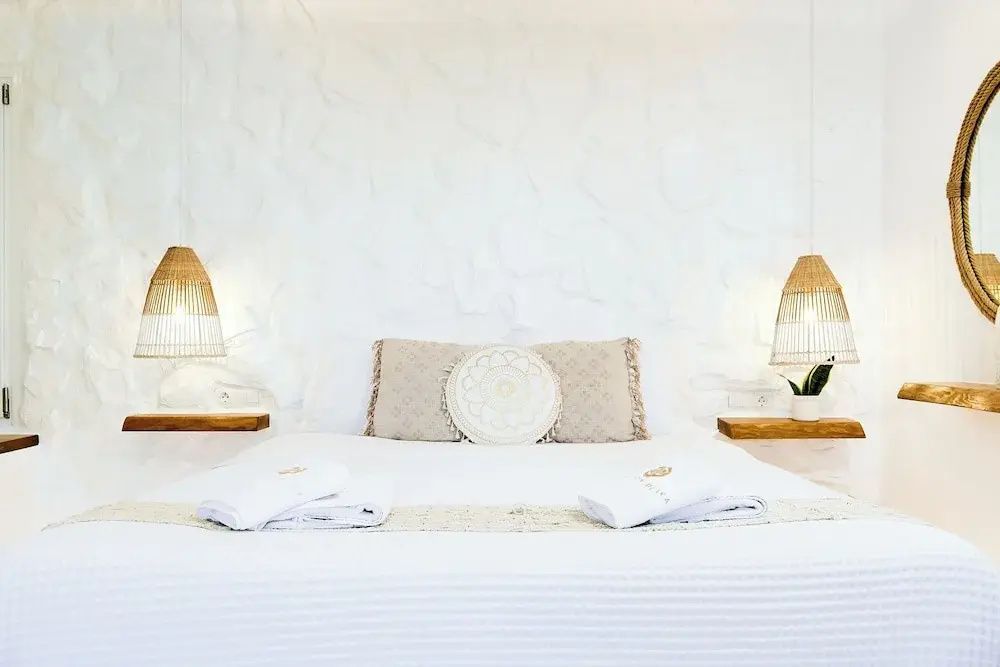 Weißes Doppelbett mit Zierkissen und Hängelampen vor einer strukturierten Wand.