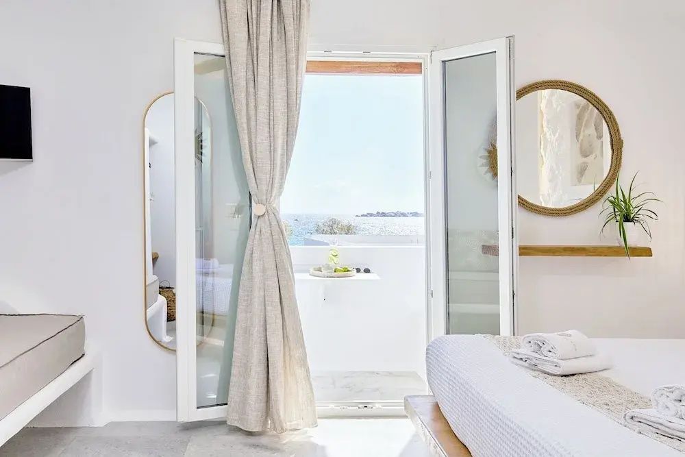 Blick durch ein geöffnetes Fenster mit hellen Vorhängen auf das blaue Meer.