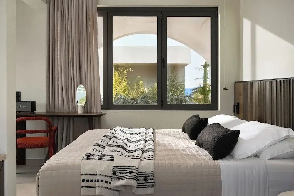 Ein Doppelbett mit hellbrauner Tagesdecke und Blick durch ein großes Fenster nach draußen.