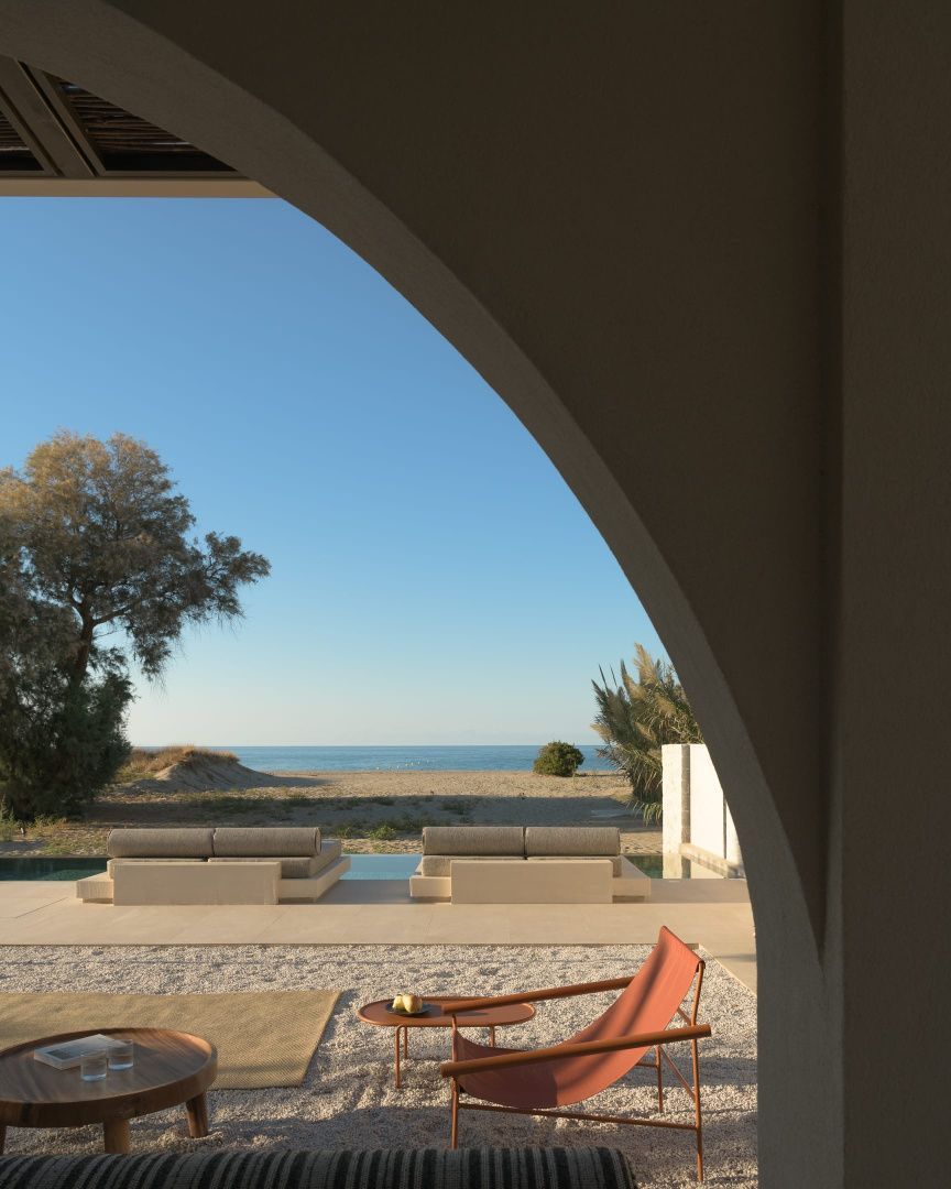 Blick durch einen Rundbogen auf eine Terrasse mit Liegen und den Sandstrand am Meer.