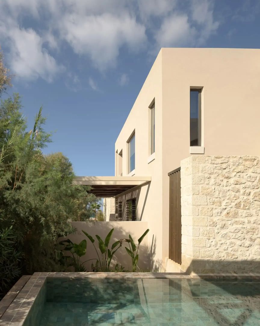 Beige Fassade eines Gebäudes mit Natursteinmauer, grünem Garten und Poolbereich bei Sonnenschein.