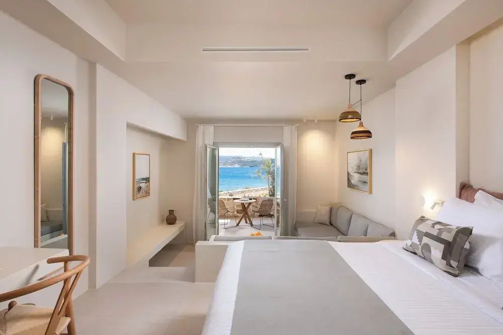 Großzügiges Zimmer mit weißem Doppelbett, Sofa und Ausblick durch die Balkontür aufs Meer.