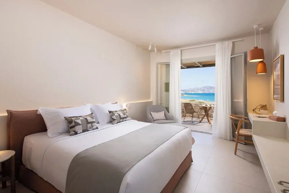 Doppelbett in hell gestaltetem Zimmer mit Blick auf die überdachte Terrasse und das Meer.