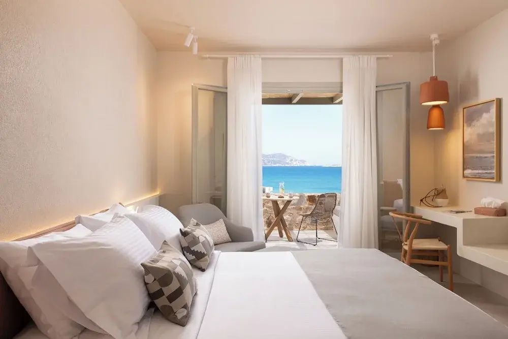 Helles Hotelzimmer mit großem Doppelbett und Blick durch die offene Balkontür aufs Meer.