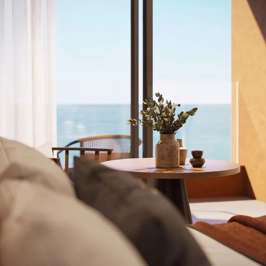 Nahaufnahme einer Vase mit Zweigen auf einem Holztisch vor einer Balkontür mit Meerblick.