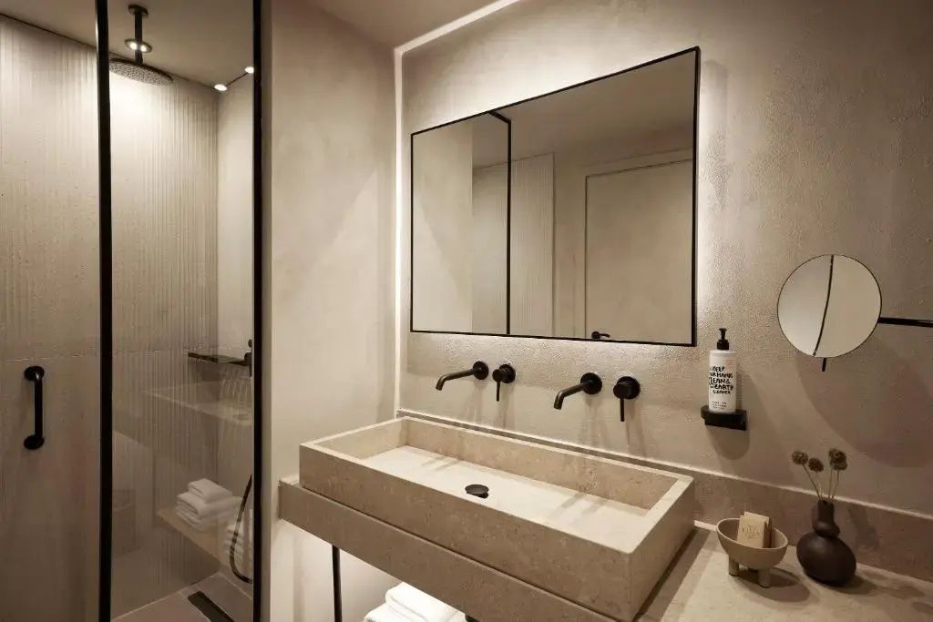 Badezimmer mit beigefarbenem Waschtisch, zwei schwarzen Armaturen und großem beleuchtetem Spiegel.