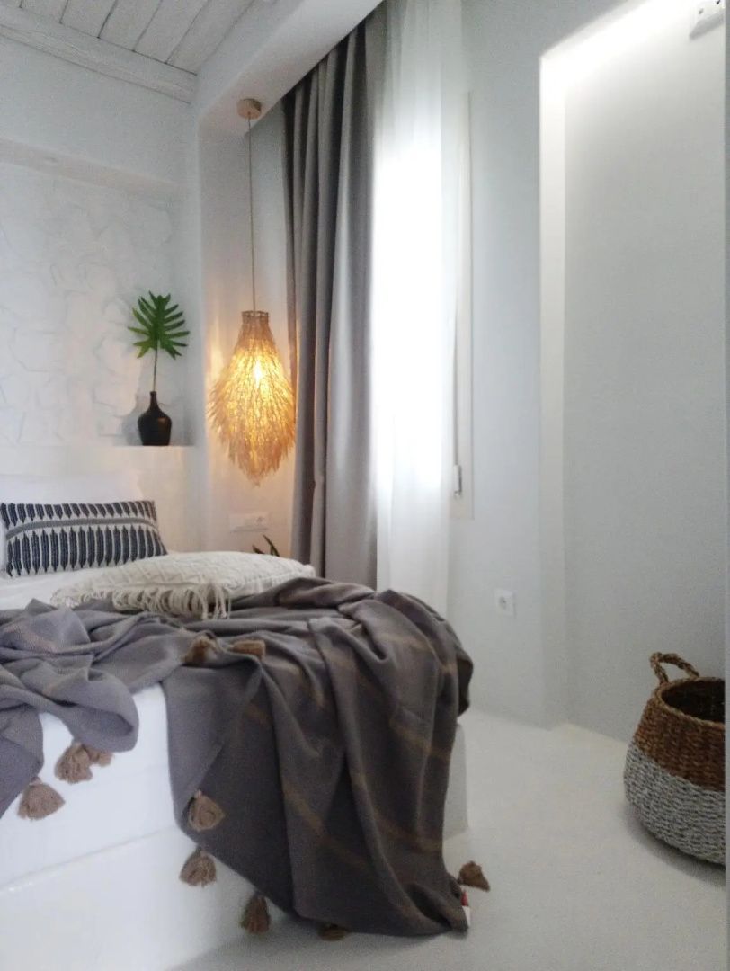 Helles Hotelzimmer mit weißem Bett, grauer Tagesdecke, geflochtener Hängelampe und Vorhang.