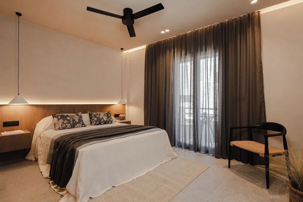 Doppelbett mit hellem Überwurf, Kissen, Deckenventilator und dunklem Vorhang am Balkon.