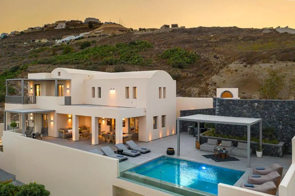 Beleuchtete weiße Villa am Abend mit blau schimmerndem Pool und Liegestühlen auf der Terrasse.