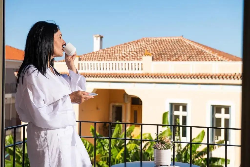 Eine Frau im weißen Bademantel trinkt Kaffee auf einem Balkon mit Blick auf Häuser.