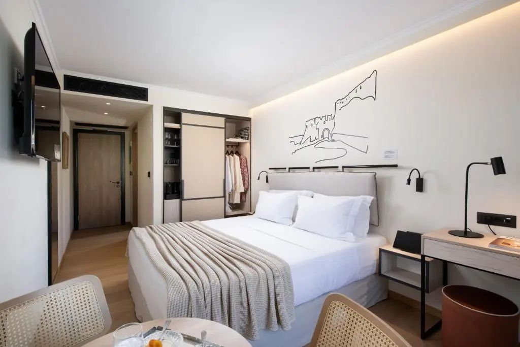 Doppelbett mit weißer Bettwäsche, heller Überwurf, Wandschmuck und offener Kleiderschrank im Hotelzimmer.