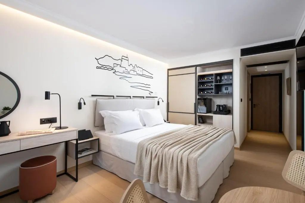 Helles Hotelzimmer mit Doppelbett, Wandzeichnung und offenem Schrank mit Minibar.