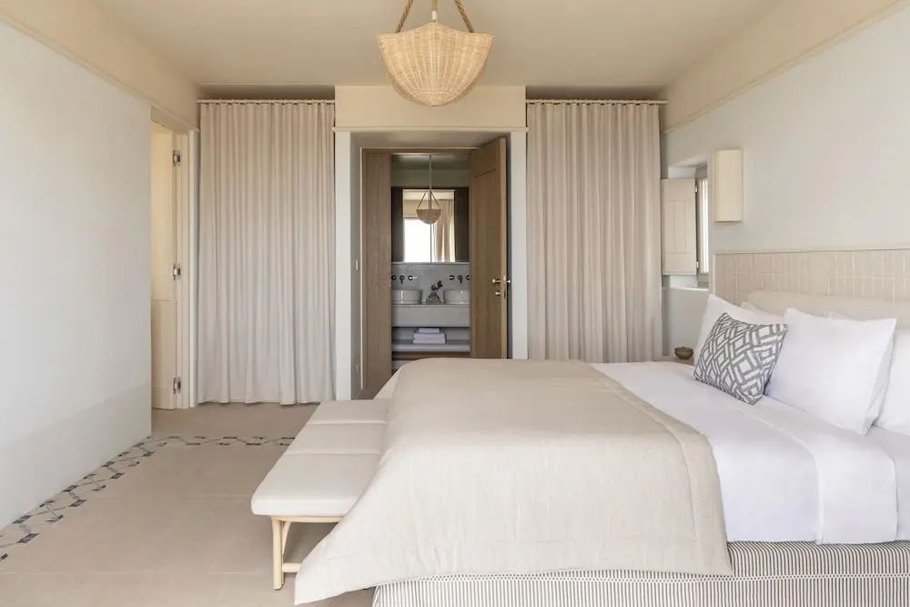 Hotelzimmer in Beigetönen mit Doppelbett, Sitzbank und Blick in das angrenzende Badezimmer.