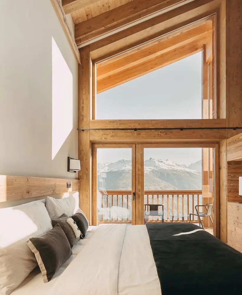 Helles Hotelzimmer aus Holz mit Doppelbett und Panoramafenster mit Blick auf die Berge.