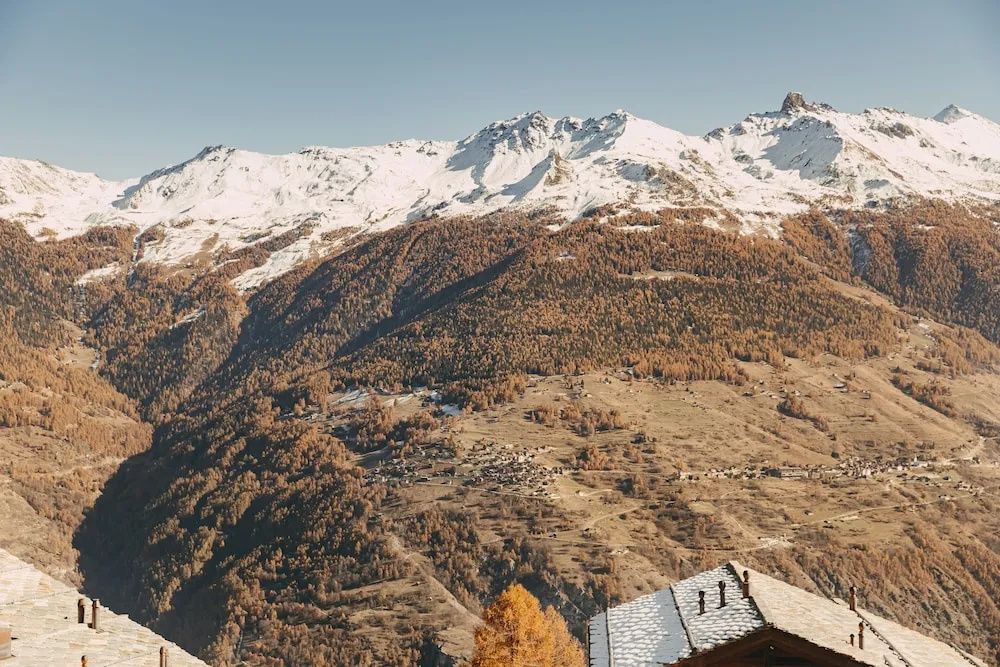 Verschneite Berggipfel und herbstliche Hänge im alpinen Tal unter blauem Himmel.