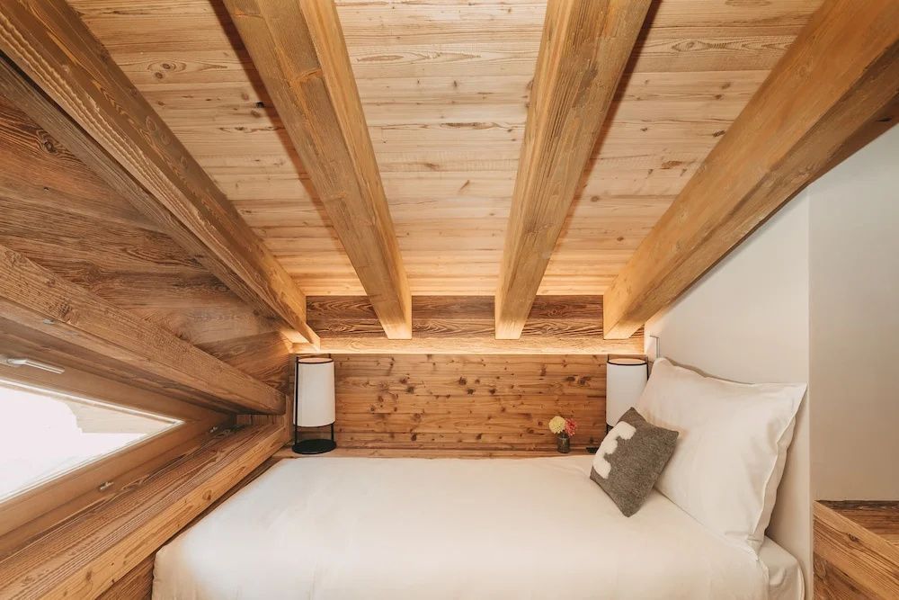 Bett unter einer schrägen Holzdecke mit freiliegenden Balken und Beleuchtung.