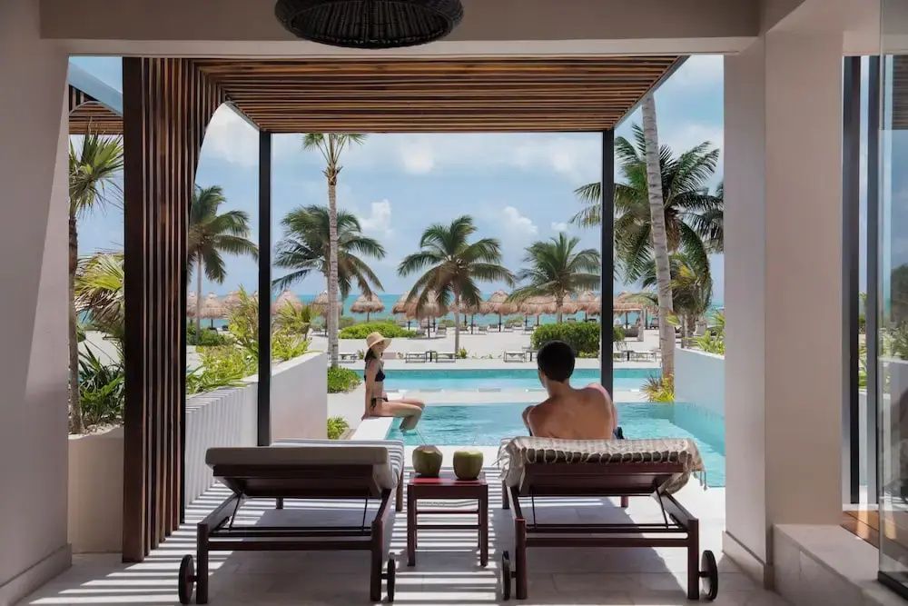 Blick von einer überdachten Terrasse mit Liegestühlen auf den Pool und Palmen am Strand.