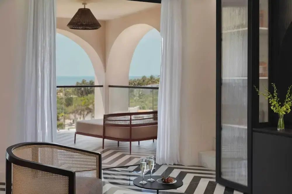 Überdachte Terrasse mit Rundbögen, Liegebett und Blick auf Palmen und das Meer.