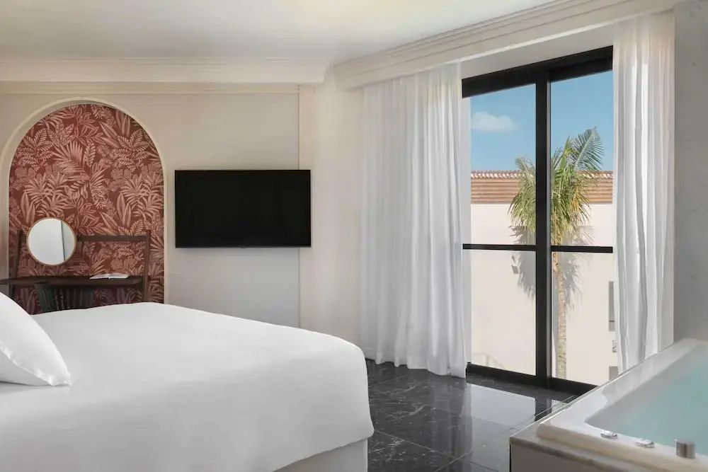 Schlafbereich mit weißem Bett, Flachbildfernseher an der Wand und großem Fenster.