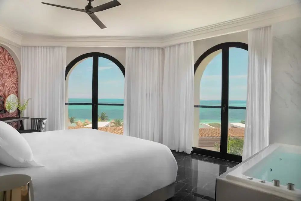 Helles Hotelzimmer mit weißem Bett und großer Fensterfront mit Meerblick.