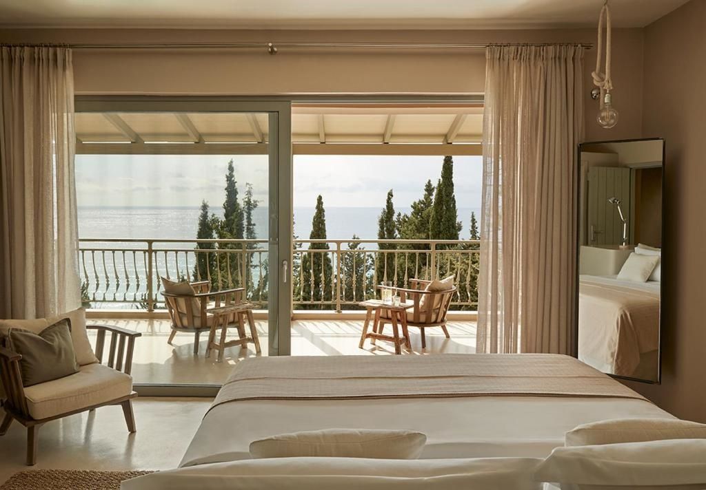 Blick aus dem Schlafzimmer auf eine Terrasse mit Holzstühlen und das Meer hinter Zypressen.