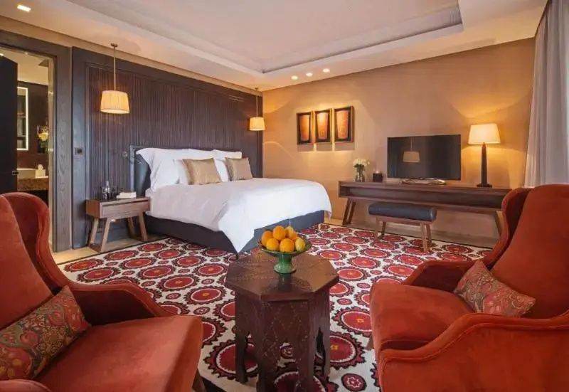 Geräumiges Hotelzimmer mit Doppelbett, rotem Teppich, Fernseher und rötlichen Sesseln.