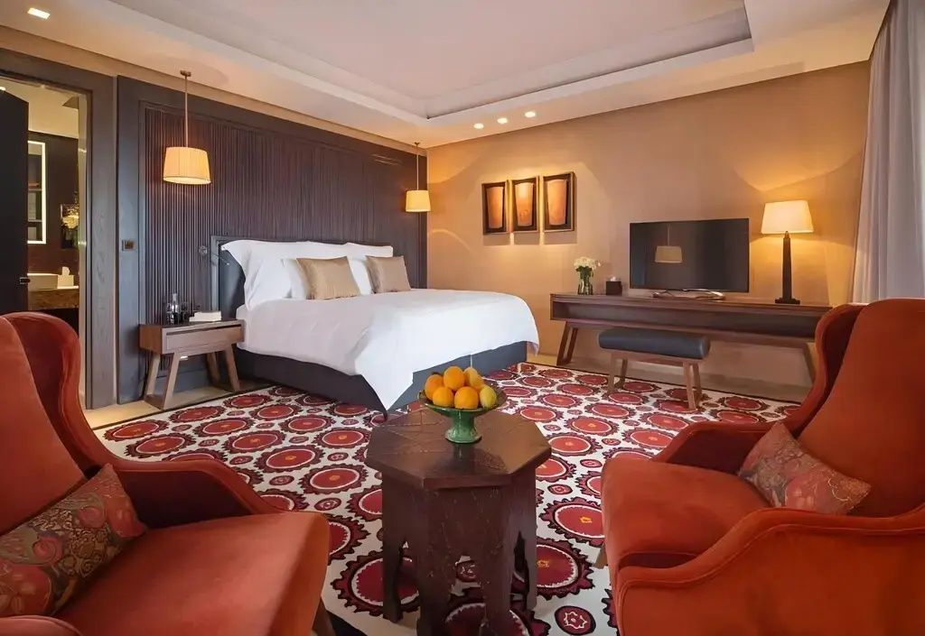 Hotelzimmer mit weiß bezogenem Doppelbett, rotem Teppich und Sitzbereich.