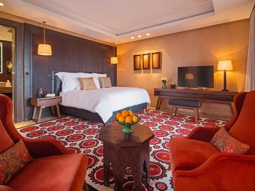 Hotelzimmer mit großem Doppelbett, rotem Teppich, Fernseher und Sesseln.