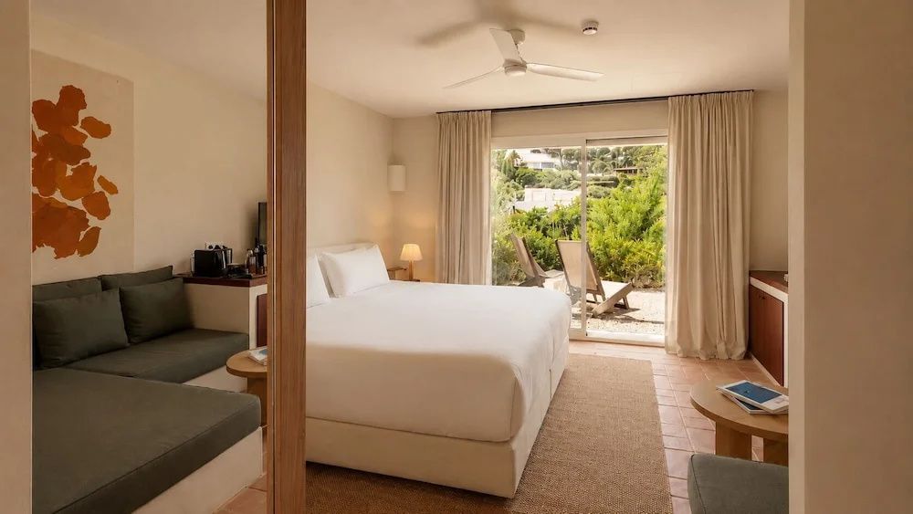 Helles Hotelzimmer mit großem Doppelbett, Deckenventilator und Zugang zur Terrasse mit Liegen.