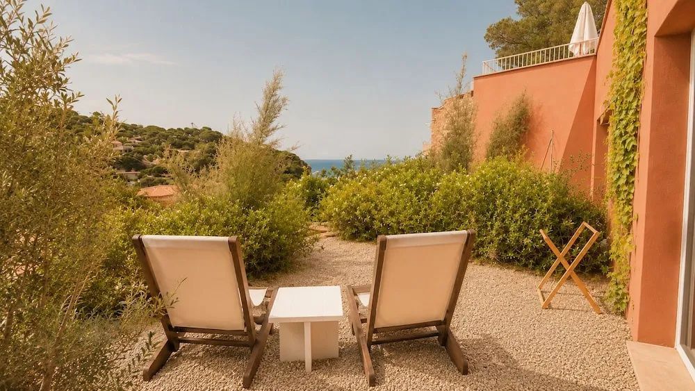 Zwei Holzliegen auf einer Terrasse mit Blick auf grüne Pflanzen und das Meer im Hintergrund.