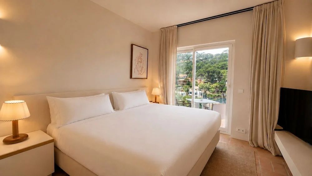 Helles Doppelzimmer mit weiß bezogenem Bett und Balkontür mit Blick ins Grüne.