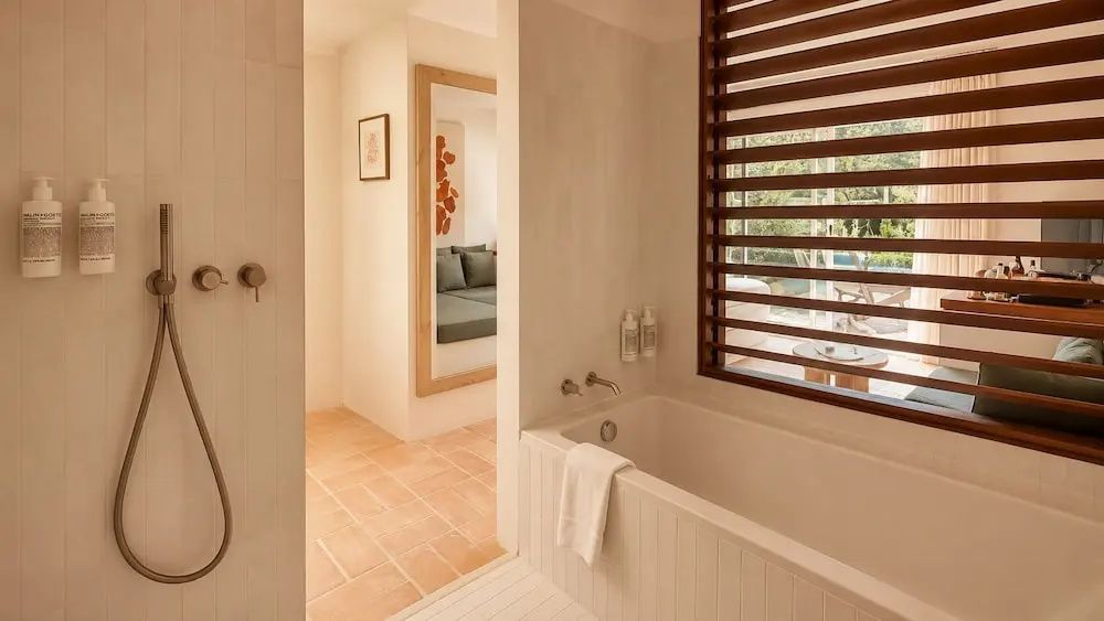 Badezimmer mit einer weißen Badewanne, Holzlamellenfenster und warmen Fliesenböden.