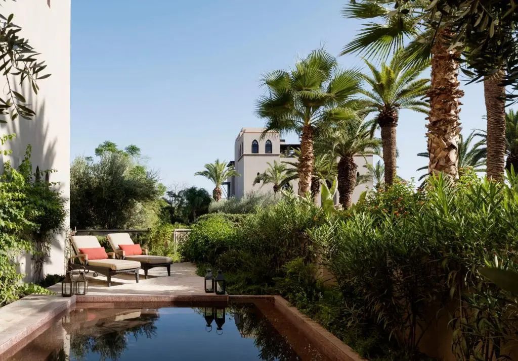 Außenpool im Gartenbereich umgeben von Palmen, Sonnenliegen und marokkanischer Architektur.