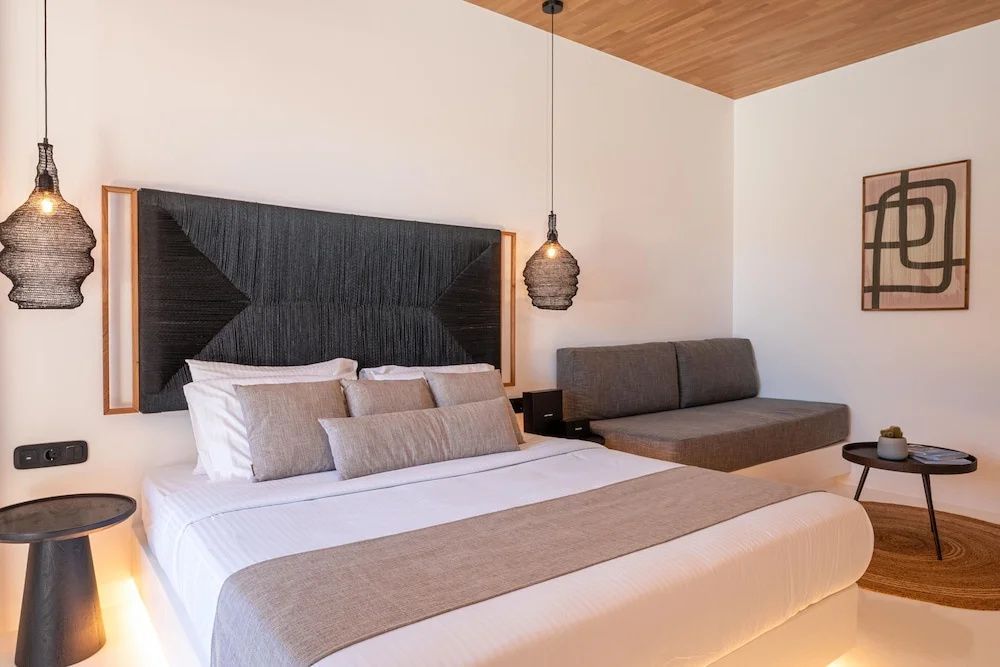 Doppelbett mit dunklem Kopfteil, Kissen, Hängeleuchte und angrenzender Sitzcouch.