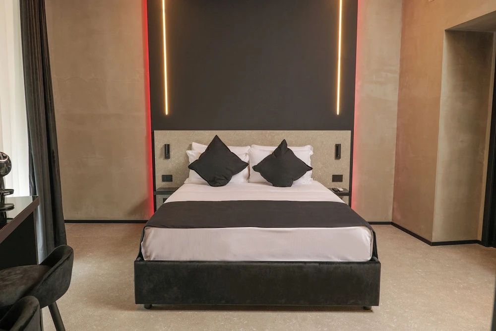 Doppelbett mit schwarzen Kissen und moderner Akzentbeleuchtung an der dunklen Wand.