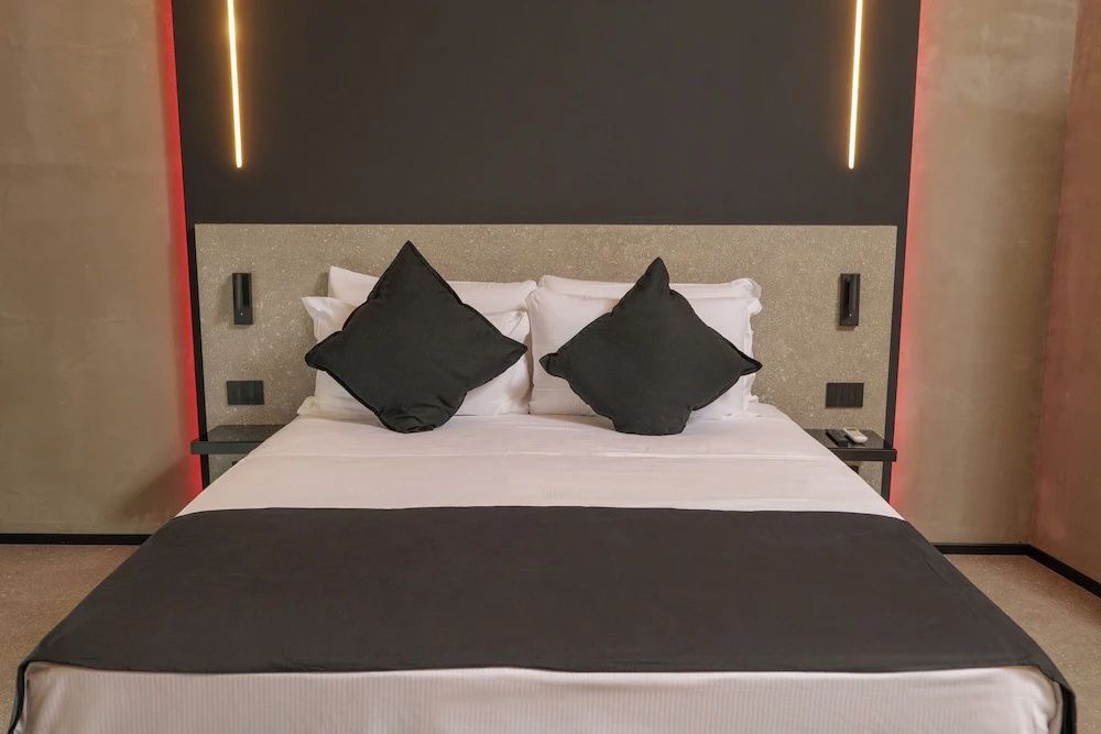 Doppelbett mit weißer Bettwäsche, zwei dunklen Kissen und moderner Wandbeleuchtung.