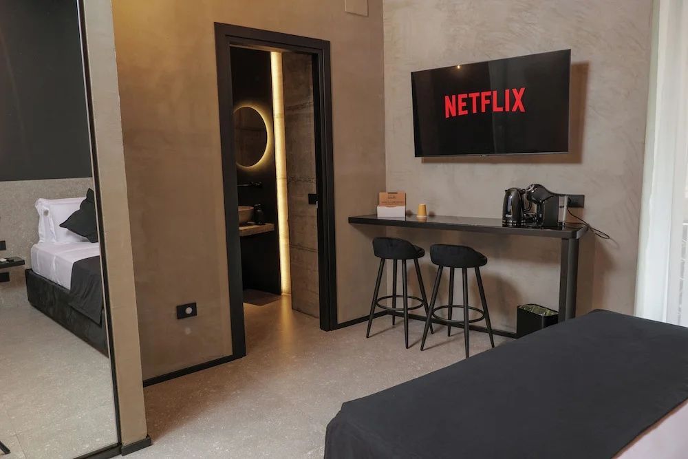 Modernes Hotelzimmer mit Flachbild-TV an der Wand, zwei Barhockern und Blick ins Badezimmer.