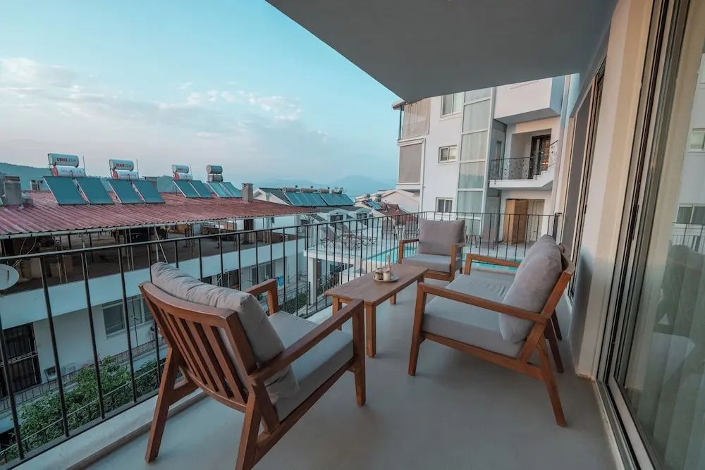 Balkon mit Holzmöbeln und grauen Sitzkissen mit Aussicht auf Nachbargebäude und Berge.