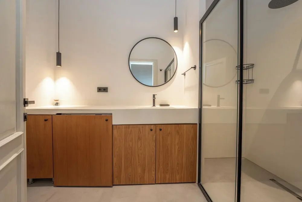 Modernes Badezimmer mit weißem Waschtisch, Holzschränken, rundem Spiegel und Glasdusche.