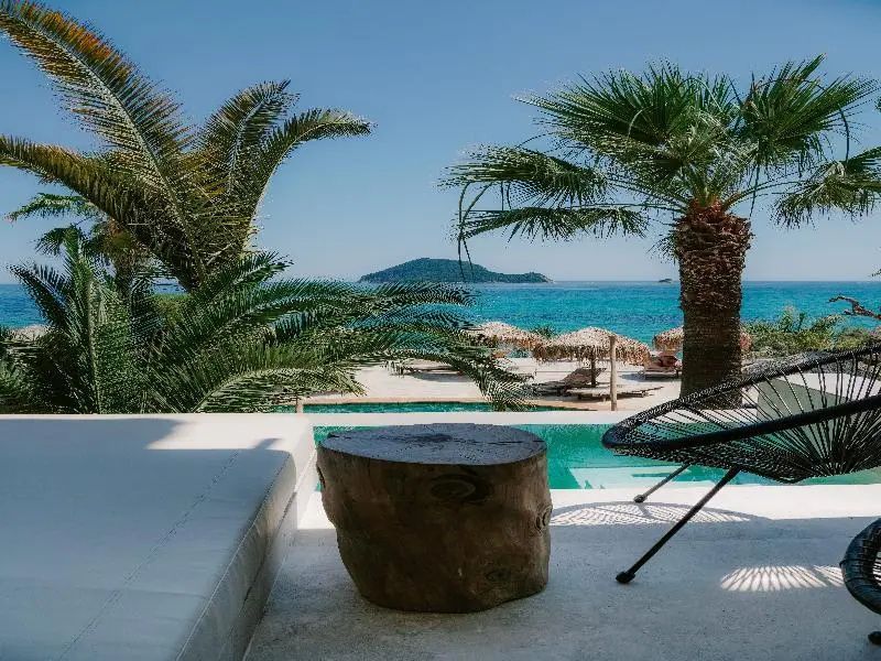 Ausblick von der Terrasse mit Holzbeistelltisch auf den Pool, Palmen und das angrenzende Meer.