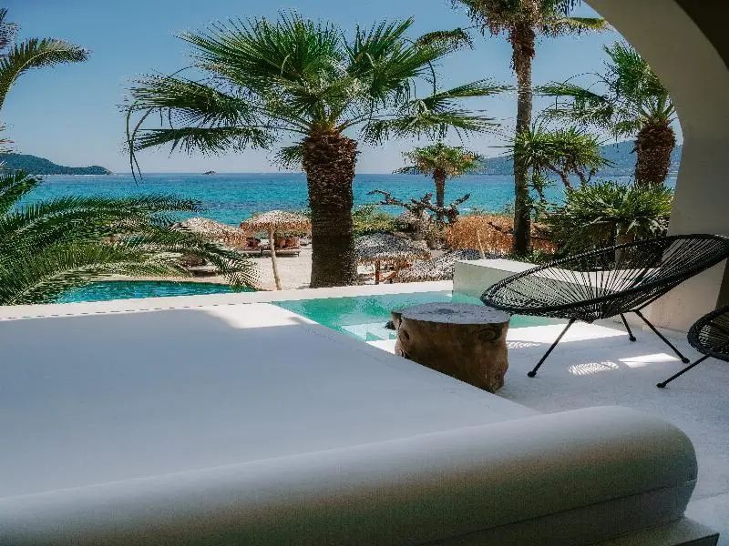 Blick von einer Terrasse mit Liegebett und Stuhl auf einen Pool, Palmen und das Meer.