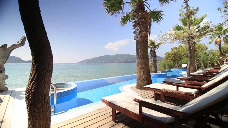 Infinity-Pool auf einer Holzterrasse mit Sonnenliegen und Blick auf das Meer und Hügel.