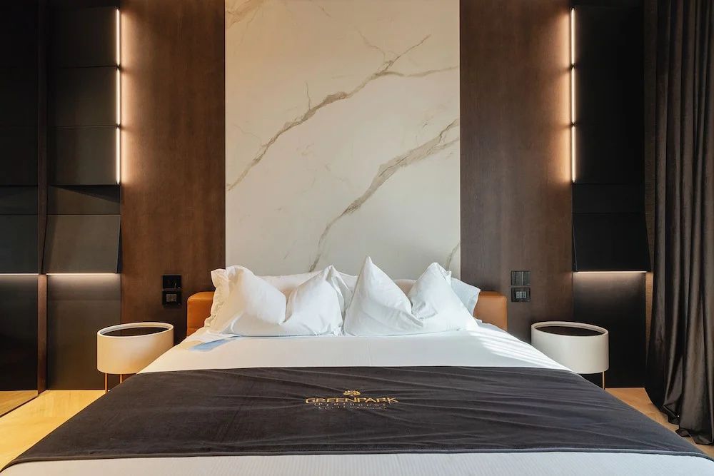 Doppelbett mit weißen Kissen, dunkler Tagesdecke und heller Marmorwand dahinter.