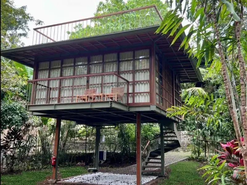 Zweigeschossige Holzvilla auf Stelzen im Grünen mit Balkon, Sitzecke und Wendeltreppe.