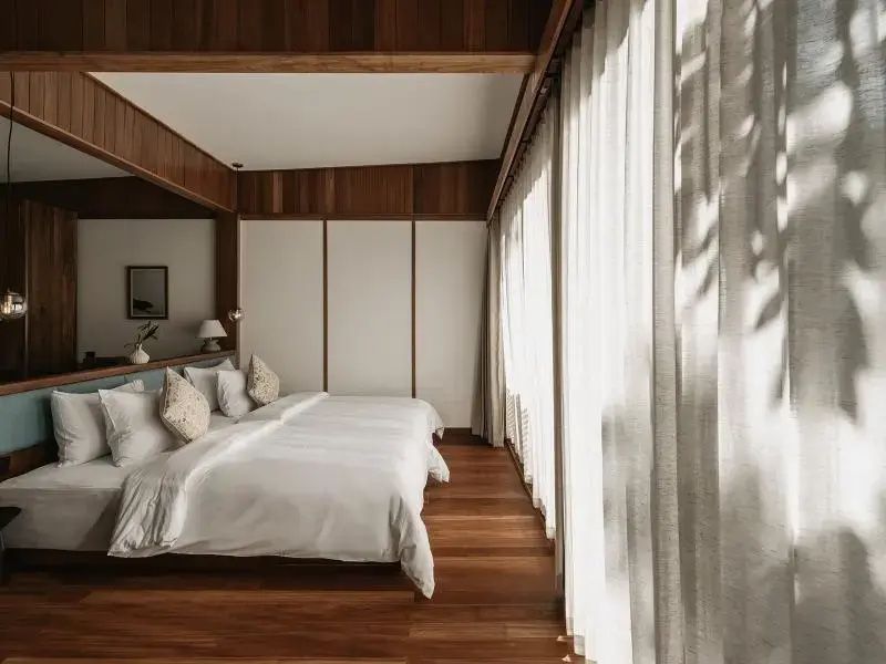 Schlafzimmer mit weiß bezogenem Bett, Holzleisten an der Decke und hellem Vorhang.