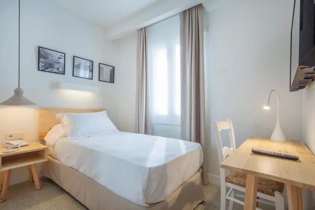 Helles Hotelzimmer mit Einzelbett, Holzschreibtisch, Stuhl und lichtdurchlässigem Vorhang am Fenster.
